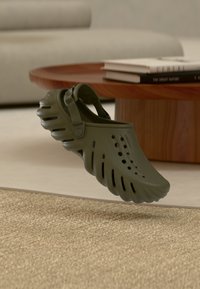 Sabot ventilé vert foncé avec une bride, flottant au-dessus d'un tapis beige dans un salon près d'une table basse en bois avec des livres.