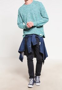 Zelený pletený svetr, tmavá džínová košile uvázaná v pase, černé džíny a modré kotníkové tenisky. Ležérní vrstvení s kontrastními barvami.