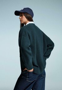 Tmavě zelený pletený svetr s texturovaným povrchem, s žebrovanými manžetami a lemem, nošený přes bílou košili s límečkem, v kombinaci s tmavými džínami.