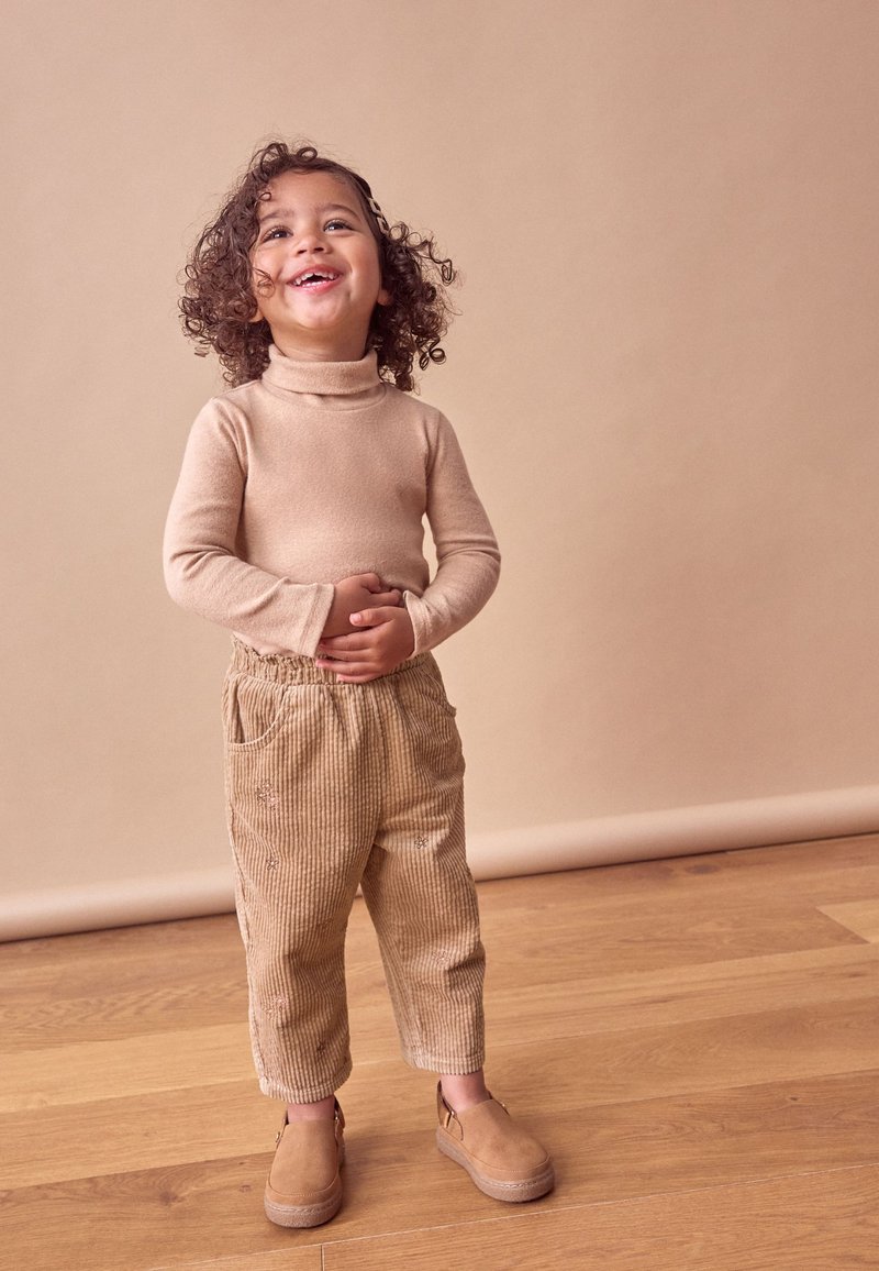 Beige turtleneck sweater, beige corduroy bukser med lommer og brune ruskindsko. Barnet står på trægulv med en neutral baggrund.