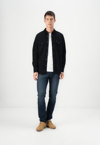 Camisa preta de botões, t-shirt branca por baixo, jeans azuis e sapatilhas bege. A camisa possui dois bolsos frontais e uma textura suave.