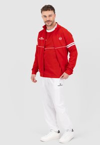 Rode sportjack met witte strepen over een rode t-shirt, gecombineerd met witte broek en witte sneakers. Logo op de kleding zichtbaar.