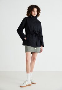 Jachetă neagră matlasată cu guler înalt, talie cu curea și buzunare laterale, asortată cu o fustă din denim verde și adidași albi.