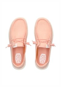 Zapatillas de lona rosa claro con una textura suave, puntera redonda y suela blanca. Presentan un diseño sin cordones y la marca "HEY DUDE" en la plantilla.