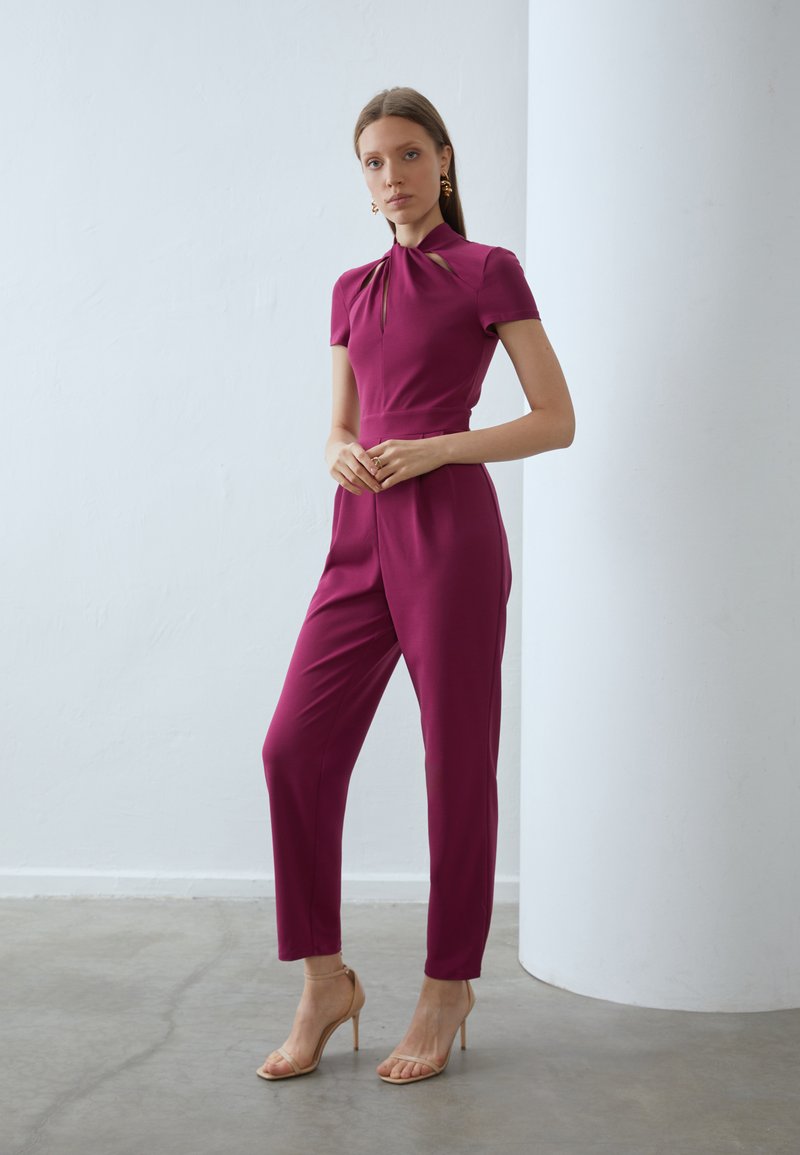 Combinaison ajustée bordeaux à manches courtes, avec un décolleté en trou de serrure et des jambes fuselées, assortie de sandales à talons hauts nudes.