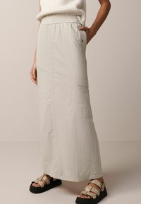 Jupe maxi gris clair en tissu texturé, avec une taille élastique, des poches latérales et une coupe décontractée. Associée à des sandales noires chunky.