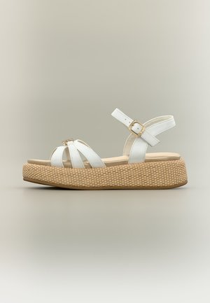 Valge rihmaga sandaal reguleeritava pahkluu pandlaga ja beežist punutud platvormpõhjaga, esitatud küljelt neutraalse tausta ees.