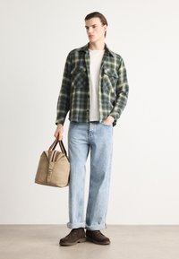Camicia di flanella a quadri verdi e blu, t-shirt bianca, jeans azzurri chiari, scarpe marroni e borsa beige con tracolle scure, in piedi davanti a uno sfondo semplice.