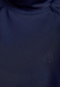 Tumesinise värviga sporditopp, valmistatud siledast kangast, millel on kõrge krae ja peen logo sarnases toonis. Õmblused on servade ümber nähtavad.