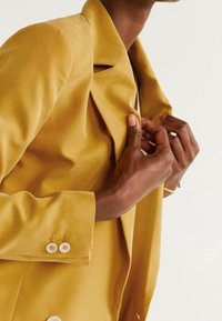 Blazer jaune moutarde avec un col structuré, un tissu lisse et des poignets à trois boutons. Dispose d'une unique découpe sur la poitrine et d'une coupe ajustée.