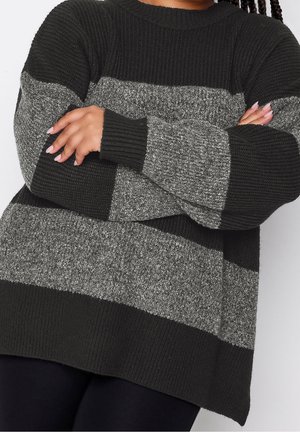 Personne croisant les bras, portant un pull en tricot oversize à rayures noires et grises avec des poignets et un ourlet côtelés.