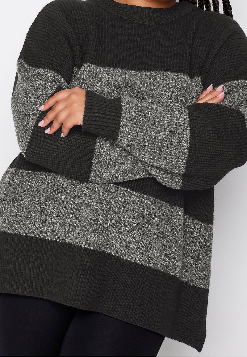 Personne croisant les bras, portant un pull en tricot oversize à rayures noires et grises avec des poignets et un ourlet côtelés.