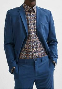 Modrý oblek z lehké látky s jedním knoflíkem, doplněný vzorovanou květinovou košilí s barevnými akcenty.