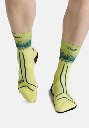 Benen met gele sneakersokken met groen gepixelde print en verticale turquoise strepen, staand tegen een witte achtergrond.