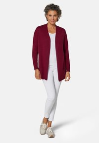 Bordeaux vest met open voorkant en lange mouwen, gecombineerd met een witte top en slanke witte broek. Lichtbruine sneakers maken de outfit af.