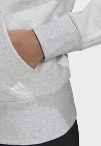 Personne portant un sweat Adidas gris clair avec la main partiellement dans la poche avant, montrant le logo sur la zone de la poche.