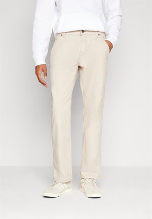 Uomo che indossa pantaloni beige chiaro a gamba dritta, maglia a maniche lunghe bianca e sneakers beige, in piedi con una mano in tasca.