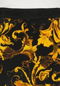 Musta kangas, jossa on koristeellinen keltainen ja ruskea kukkakuosi. Sisältää tekstuuritetun vyötärönauhan, jossa on kohokuvioitu "VERSACE JEANS COUTURE".