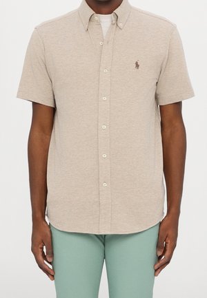 Homme portant une chemise beige à manches courtes boutonnée par-dessus un maillot blanc et un pantalon vert clair, debout devant un fond uni.