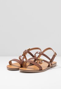 Sandalias de cuero marrón con múltiples tiras, suelas planas y un diseño minimalista. Cuentan con un cierre de hebilla y una plantilla color canela.