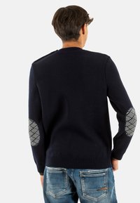 Maglione lavorato a maglia navy con scollatura rotonda, tessuto testurizzato e toppe ai gomiti con motivo. Abbinato a jeans blu scoloriti.