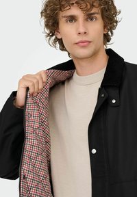 Ung mand med krøllet hår iført beige sweater og sort jakke, der viser en rød og beige mønstret quilted foring.
