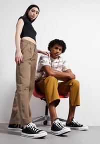 Top nero aderente, pantaloni larghi khaki, camicia a righe con maniche corte, pantaloni cropped color senape e sneakers nere indossati da due modelli su uno sfondo bianco.