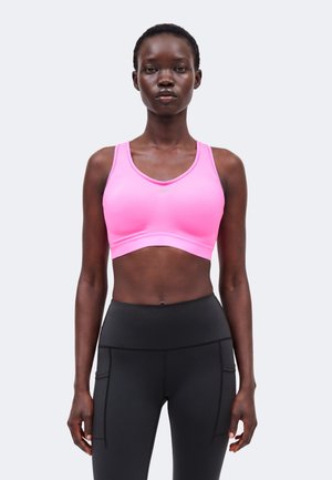 Λεπτό άτομο φοράει ένα φωτεινό ροζ αθλητικό σουτιέν και μαύρο ψηλόμεσο κολάν, στέκεται μπροστά σε ένα απλό φωτεινό φόντο.