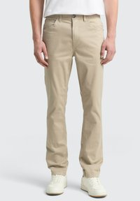 Beige Freizeithosen aus Baumwolle mit geradem Bein, fünf Taschen, Knopfverschluss und getragen mit weißen Sneakers.