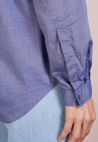 Camisa de mangas largas de color azul claro con un tejido texturizado, puños abotonados y dobladillo curvado. Combinada con jeans de mezclilla azul claro.