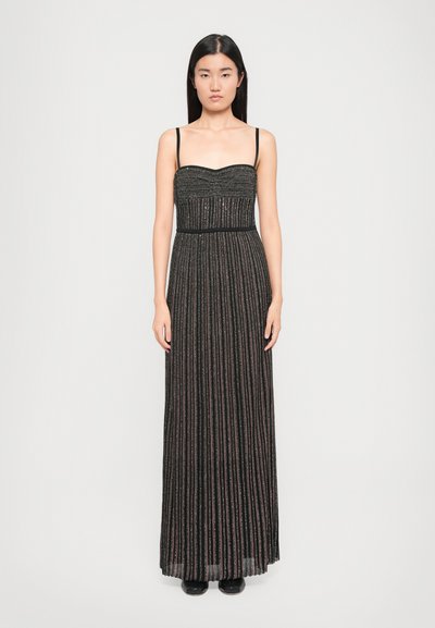 Femme se tenant de face, portant une robe plissée sans manches, noire et à rayures métalliques, de longueur maxi avec de fines bretelles et des chaussures noires.