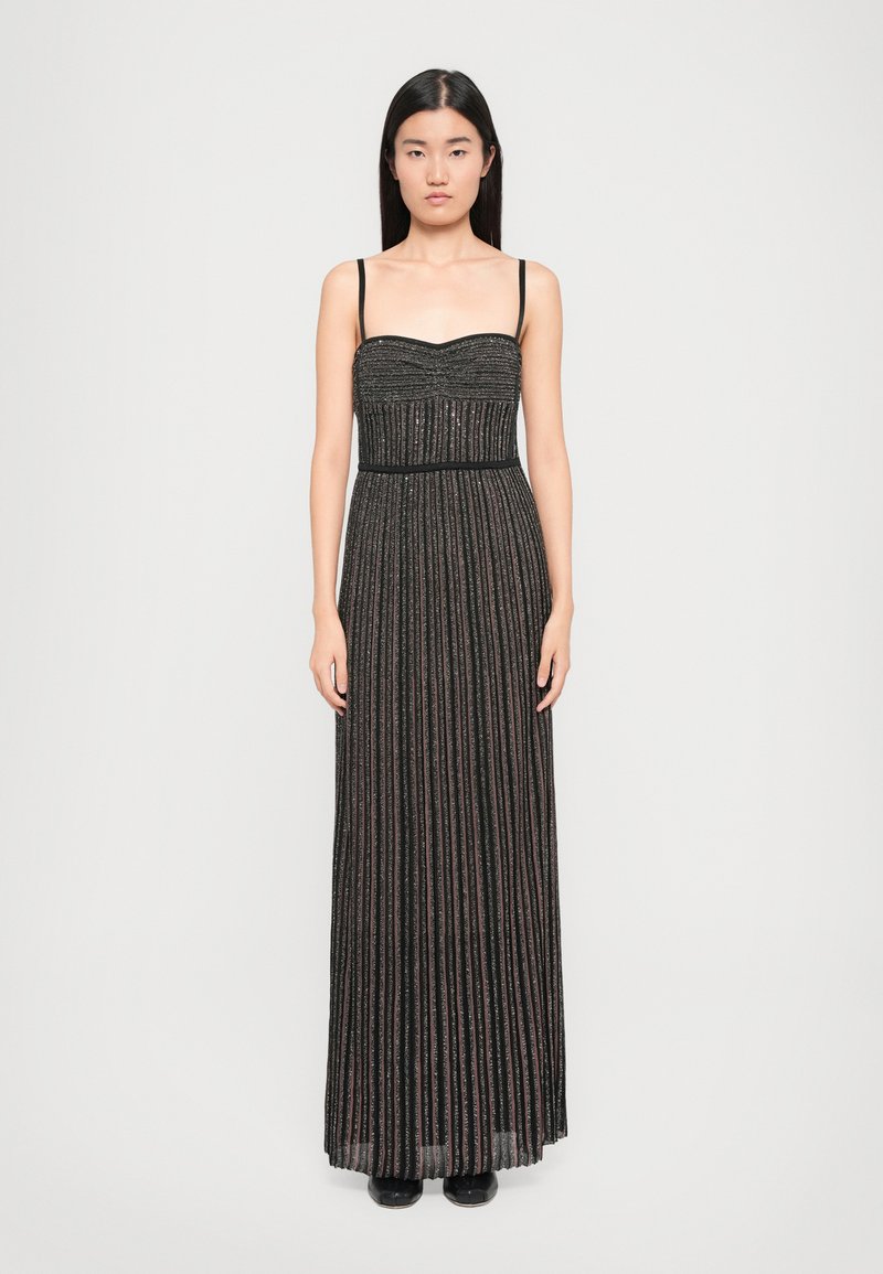 Woman stands facing forward wearing a sleeveless, black and metallic striped floor-length pleated dress with thin straps and black shoes.