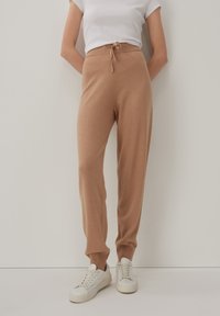 Beige gestrickte Jogginghosen mit einem elastischen Bund und Kordelzug, ausgestattet mit gerippten Beinbündchen; kombiniert mit weißen Sneakers.