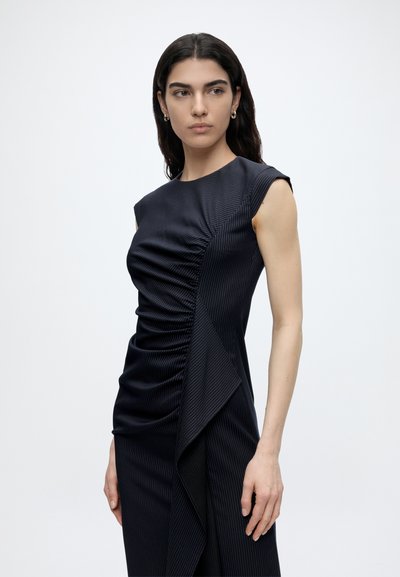 Femme aux cheveux foncés porte une robe sans manches bleu marine à fines rayures froncée avec un détail drapé asymétrique, regardant vers sa gauche sur fond blanc.