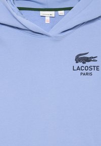 Vaaleansininen huppari, joka on valmistettu puuvillasekoitteesta. Hupparissa on kengurutasku, nyöritettävä huppu ja musta Lacoste Paris -logo rinnassa.