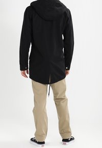 Giacca nera con cappuccio e un lungo retro, abbinata a pantaloni beige e sneakers nere, con un design minimalista e sottili dettagli in metallo.