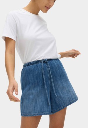 Kvinde iført hvid t-shirt med korte ærmer og blå, højtaljede denimshorts med elastisk talje og bindebånd foran, stående mod en ensfarvet baggrund.