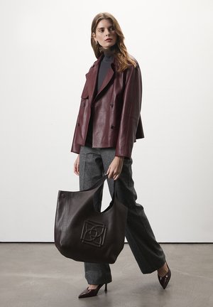 Sac en cuir marron foncé avec un design géométrique embossé, associé à une veste en cuir bordeaux, un pantalon gris et des talons aiguilles.