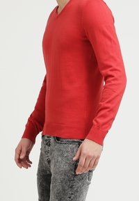 Pull en laine rouge avec un décolleté en V, des manches longues et un ourlet côtelé. Associé à un jean gris texturé, mettant en valeur un design ajusté.