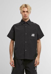 Camisa negra de botones con mangas cortas, un bolsillo con parche que lleva un logo impreso, y rayas blancas a los lados de los hombros. Textura de tela suave.