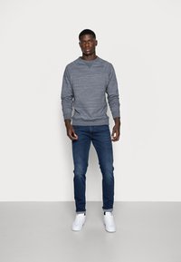Grijze sweatshirt met lange mouwen en geribbelde manchetten, gecombineerd met strakke donkerblauwe jeans en witte sneakers op een neutrale achtergrond.