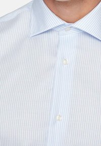 Uomo che indossa una camicia a righe blu chiaro e bianca con colletto, mostrando la parte superiore del petto e del collo.