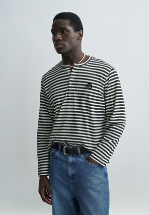 Homme portant une chemise à manches longues rayée noir et blanc avec des boutons, un jean bleu, une ceinture noire et un collier de perles, debout avec une main dans la poche.