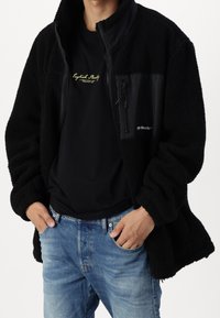 Schwarze Fleecejacke mit einer glatten, schwarzen Fläche und einer Reißverschlusstasche auf der Brust. Getragen über einem schwarzen T-Shirt und blauen Jeans.