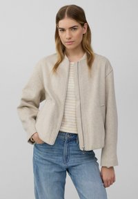 Giacca cropped beige chiaro con zip frontale, caratterizzata da un collo rotondo e maniche leggermente oversize, abbinata a un top a righe e jeans in denim.