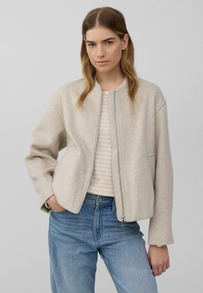 Giacca cropped beige chiaro con zip frontale, caratterizzata da un collo rotondo e maniche leggermente oversize, abbinata a un top a righe e jeans in denim.
