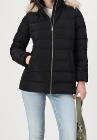 Zwarte donsjas met rits, voorzien van een met bont gevoerde capuchon, doorgestikt ontwerp en zijzakken. Gecombineerd met lichtblauwe spijkerbroek en een groene tas.