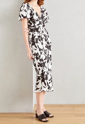 Kvinde iført en hvid midi-kjole med sort blomsterprint, flagrende ærmer og bindebånd i taljen, kombineret med sorte åbne block heel sandals.