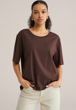 Femme aux cheveux bouclés portant une chemise ample à manches courtes marron foncé et un pantalon beige clair, les mains dans les poches, sur un fond uni.