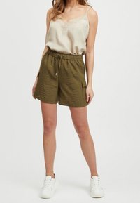 Camisola in raso beige chiaro con pizzo e pantaloni corti verde oliva con texture, dotati di una cintura elastica e tasche laterali, abbinati a sneakers bianche.